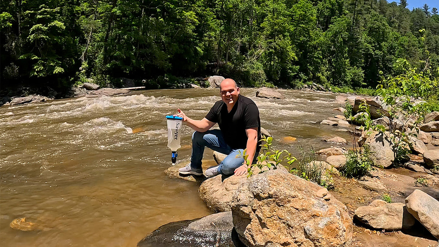 Man using Sawyer water filter by a river in mountainous terrain, showcasing outdoor survival gear.