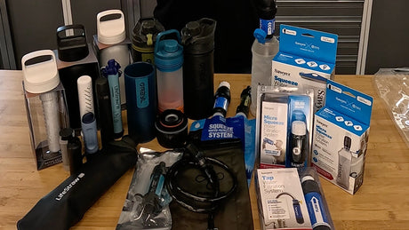Emergency water filtration gear including filter bottles, gravity systems, and hydration packs displayed on a rocky outdoor surface, ready for survival use.
